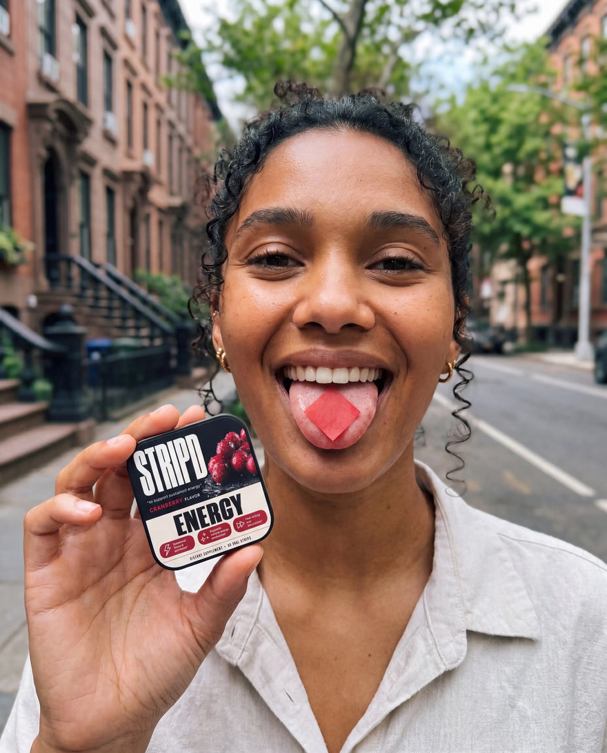 Woman smiling with a STRIPD Energy strip dissolving on her tongue, holding the cranberry Energy tin on a New York street